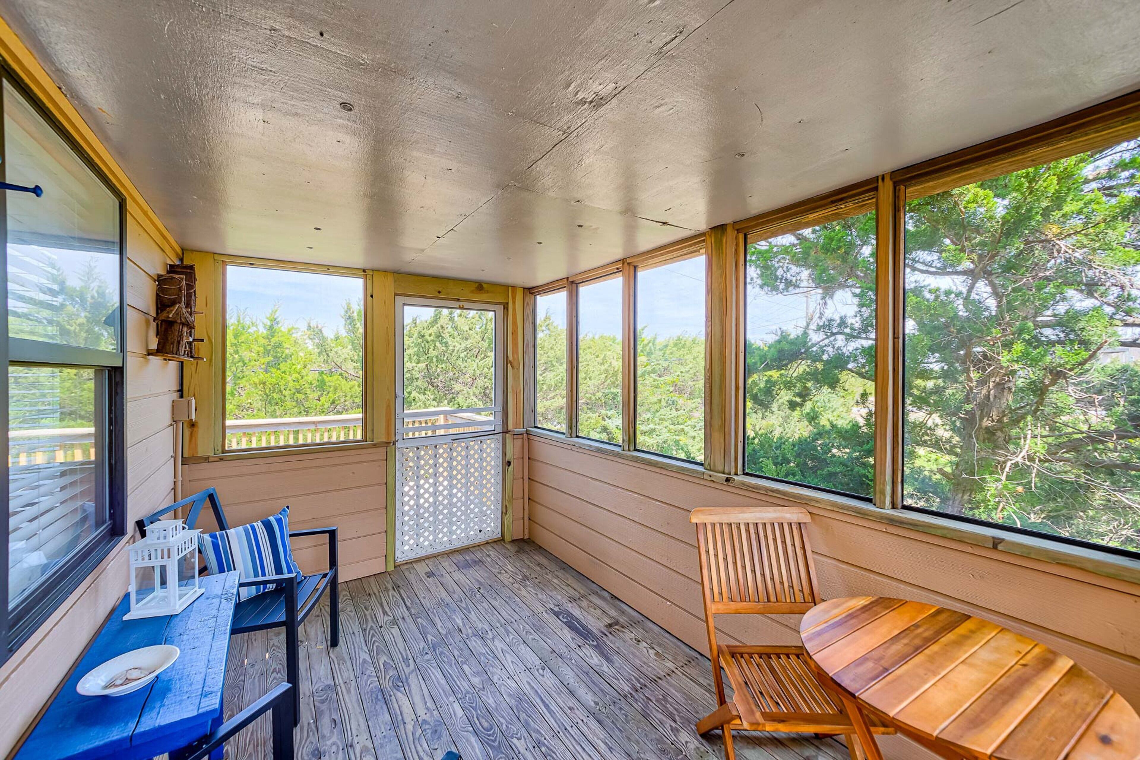 A covered porch with a wooden table and chairs, large windows, and a view of trees.