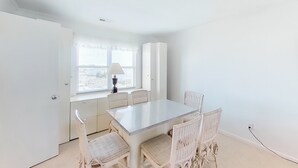 A bright, white room with a large dining table and chairs, a window with blinds, and a lamp on the windowsill. - Stunning, Multi-Level Home Steps From Beach w/WiFi, Central AC, W/D, & Patio (Holden Beach)