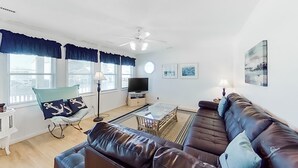A living room with a brown leather sofa, a rocking chair with nautical pillows, a glass coffee table, a flat-screen TV, and a ceiling fan. - Stunning, Multi-Level Home Steps From Beach w/WiFi, Central AC, W/D, & Patio (Holden Beach)