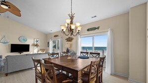 A dining area with a wooden table and chairs, a chandelier, and a view of the ocean through large windows. - Oceanfront home with great views, central AC, washer/dryer, beach access (North Topsail Beach)