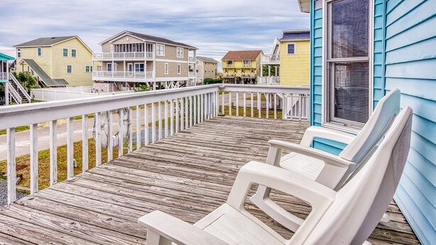 A wooden deck with white chairs and a railing, overlooking a row of colorful houses. - Fantastic house with WiFi, central AC, and private washer/dryer - steps to beach (Holden Beach)