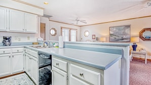 A kitchen with white cabinets, a dishwasher, and a large island with drawers. There are two windows with blinds, a ceiling fan, and a painting on the wall. - Fantastic house with WiFi, central AC, and private washer/dryer - steps to beach (Holden Beach)