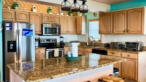 A kitchen with granite countertops, wooden cabinets, a stainless steel refrigerator, a microwave, a stove, and a dishwasher. There are two bar stools and a window with blinds. - Oceanfront Retreat, Stunning Views, Renovated Spaces, Game Room with Pool Table (Avon)