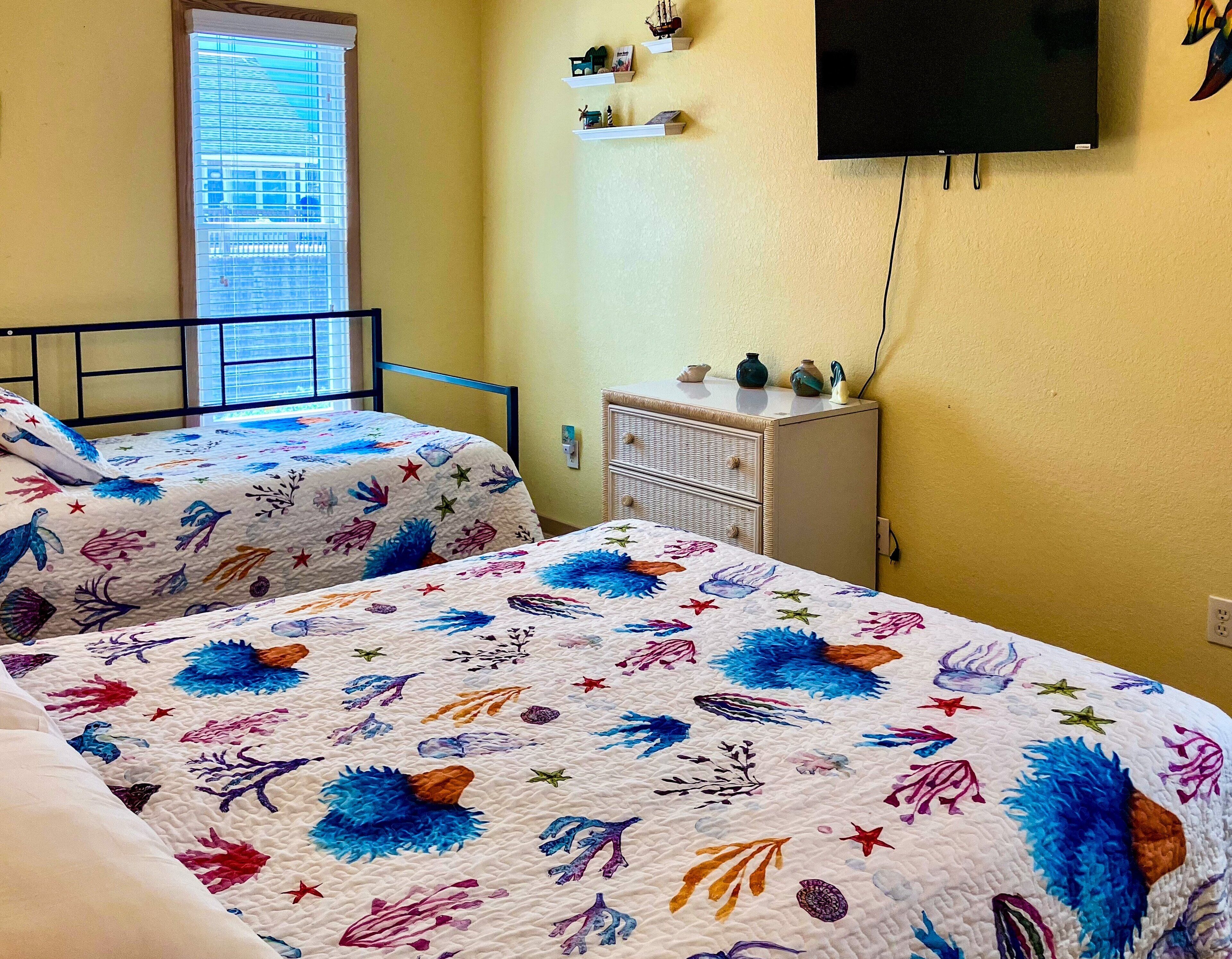 A room with two beds, each with a colorful quilt, a small dresser, a television on the wall, and a window with blinds.