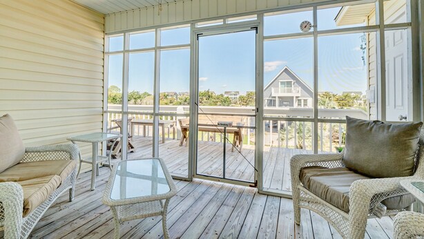 A covered outdoor deck with wicker furniture, a glass table, and a view of a house in the background. - Magnificent house with central AC, patio, deck, W/D & screened porch (Holden Beach)