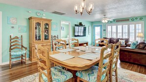 A dining area with a wooden table and chairs, a chandelier, and a television in the background. - Magnificent house with central AC, patio, deck, W/D & screened porch (Holden Beach)