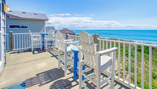 Um alpendre à beira-mar com cadeiras Adirondack brancas e uma mesa azul, com vista para o oceano.