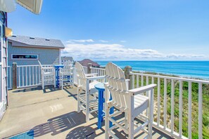 Eine Terrasse am Strand mit weißen Adirondack-Stühlen und einem blauen Tisch, mit Blick auf das Meer.