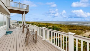 Une terrasse en bois avec des rampes blanches, du mobilier d’extérieur et une vue sur la plage et l’océan.