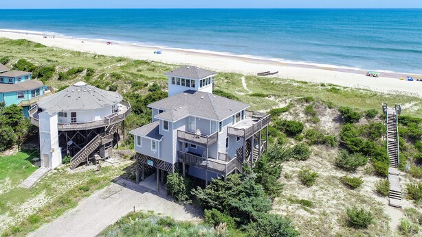 Une propriété située sur la côte, avec une grande maison à plusieurs niveaux et une tour, donnant sur une plage de sable et offrant une vue sur l’océan.