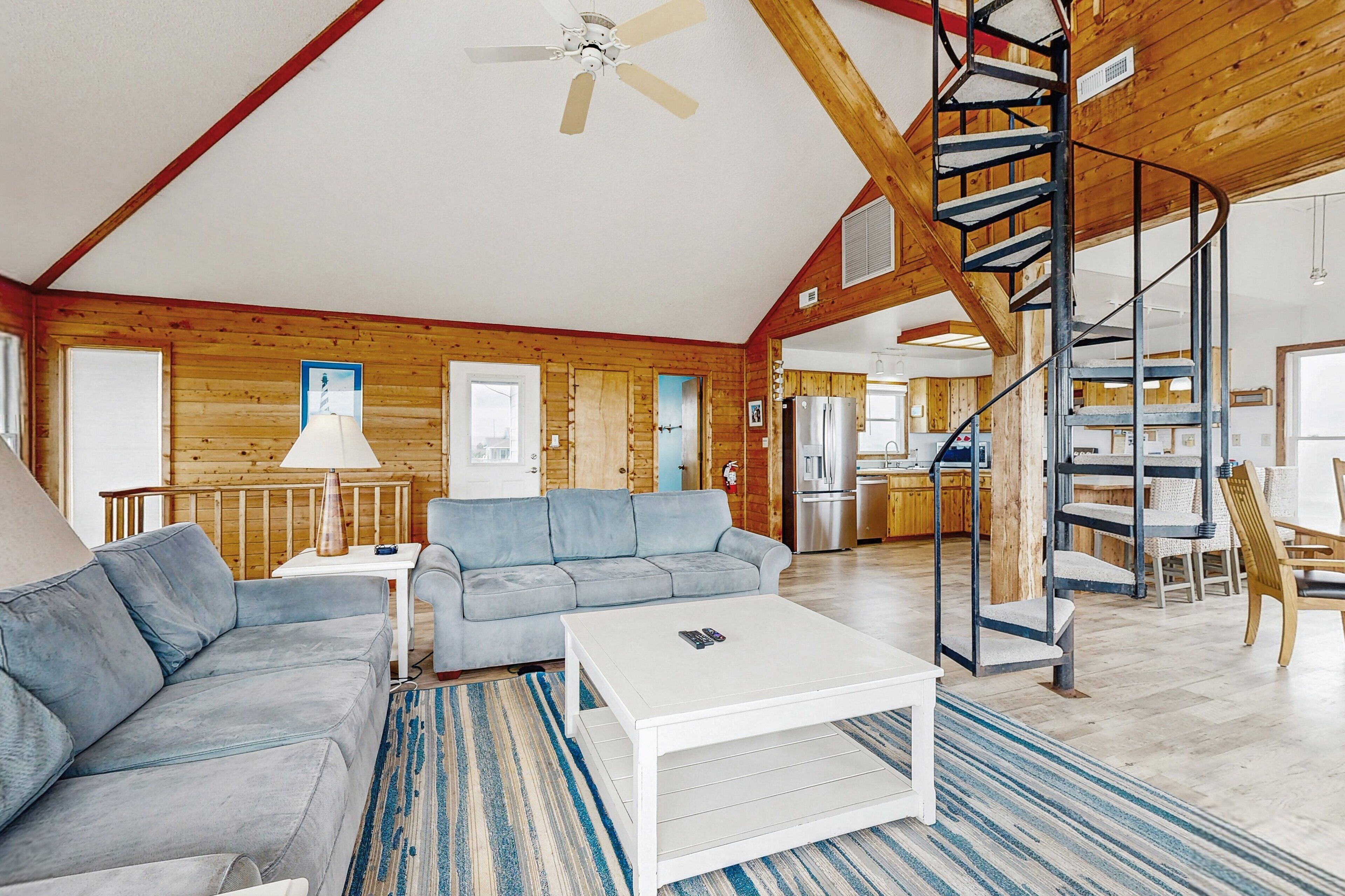 A spacious living room with a modern sofa, a white coffee table, and a blue striped rug. A spiral staircase leads to an upper level, and a kitchen is visible in the background.