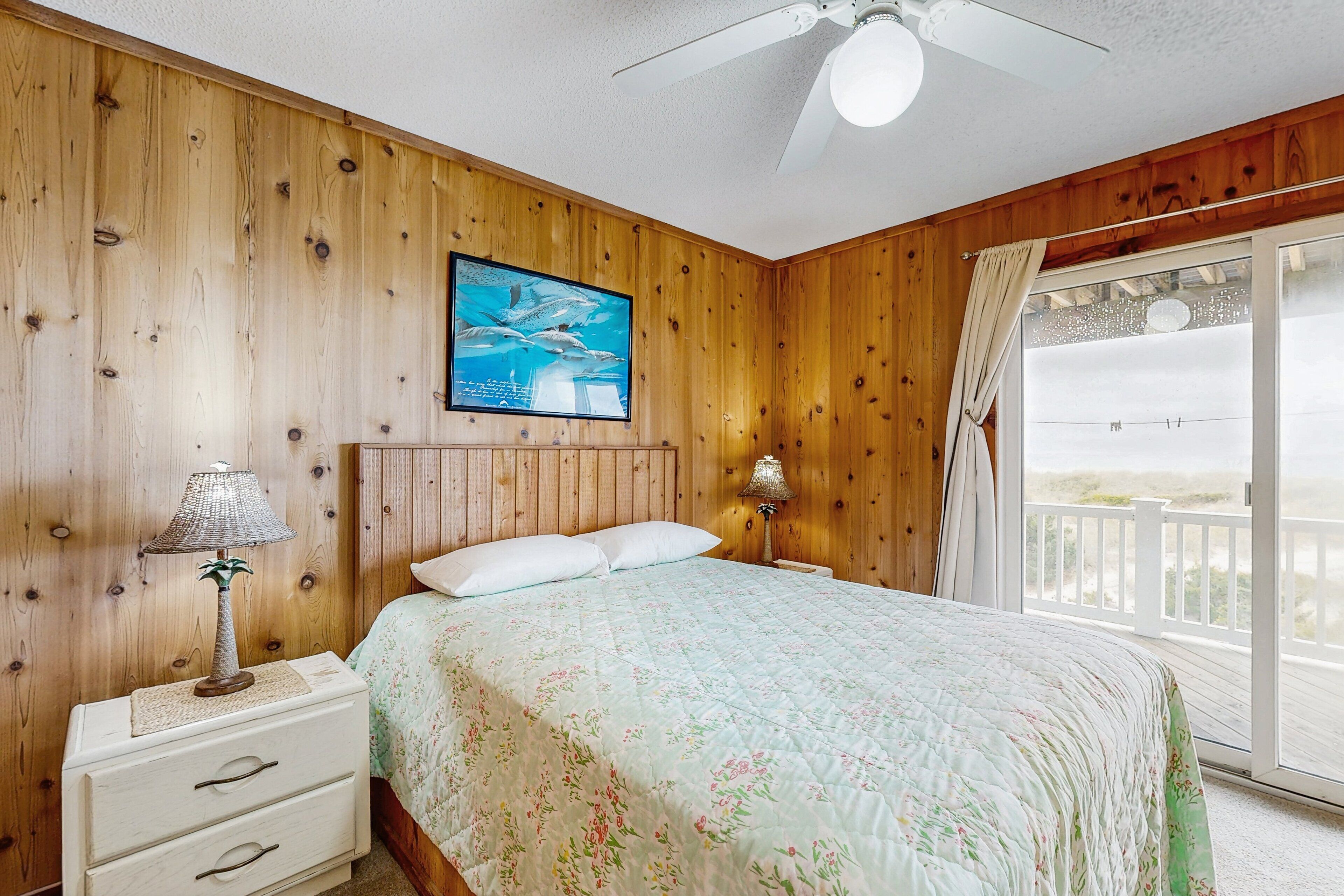 A bedroom with wooden walls, a bed with floral bedding, a ceiling fan, and a view of the beach through a large window.