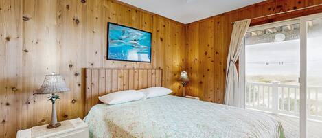 A bedroom with wooden walls, a bed with floral bedding, a ceiling fan, and a view of the beach through a large window.