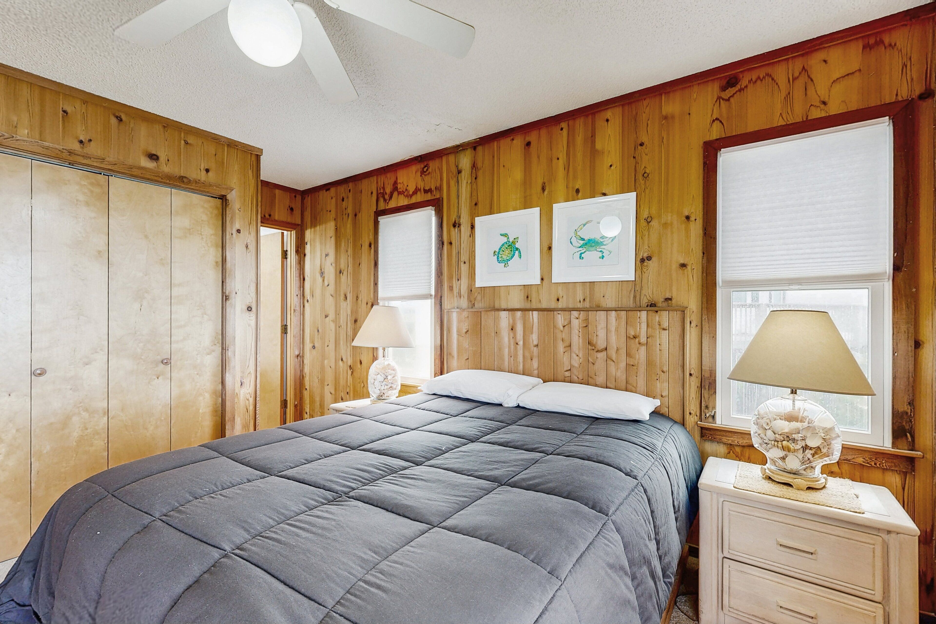 A bedroom with a bed, wooden walls, a ceiling fan, and two windows with blinds.
