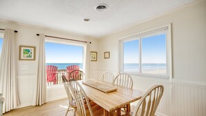 Un coin repas avec une table en bois et des chaises blanches, de grandes fenêtres équipées de stores et une vue sur l’océan.
