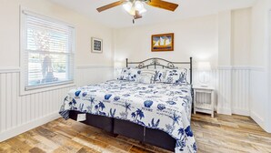Una camera da letto con pavimento in legno, un letto con copriletto a motivi blu e bianchi, una ventola a soffitto, una finestra con persiane e un comodino con lampada.