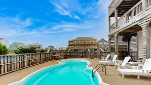 A swimming pool with a wooden deck and lounge chairs, surrounded by a wooden fence and houses in the background. - Inviting house with central AC, private pool, hot tub, ocean views, & W/D (Hatteras)