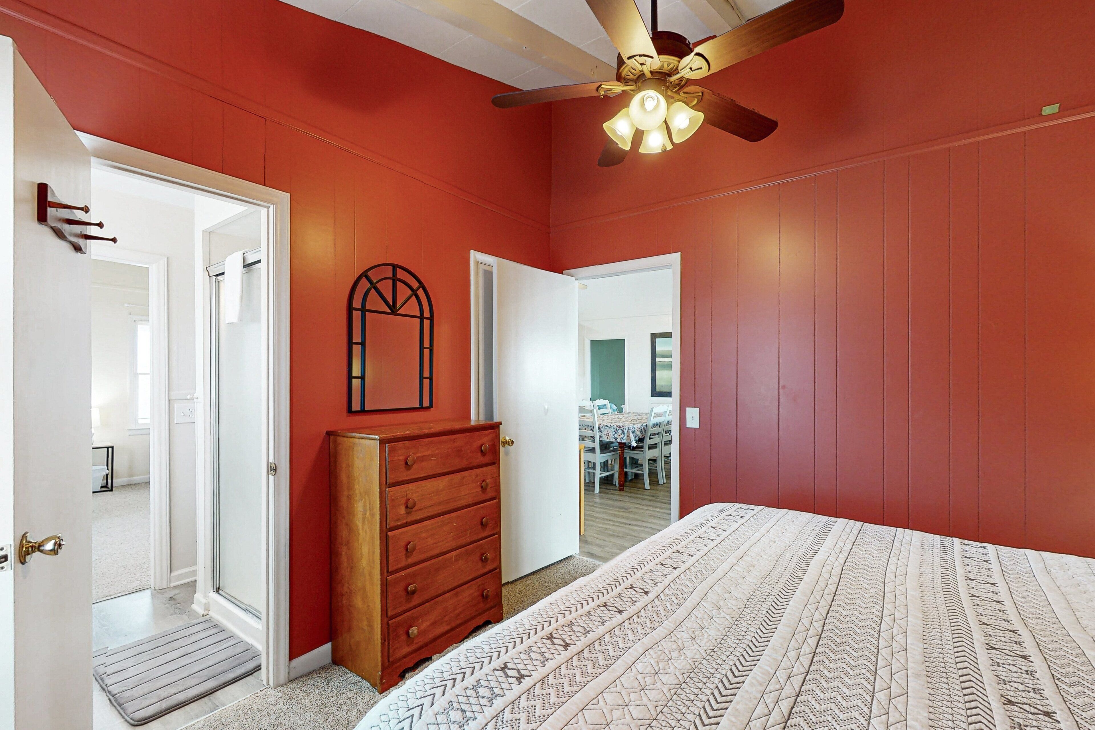 A bedroom with a bed, a dresser, a ceiling fan, and a door leading to another room.