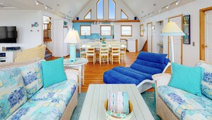 A spacious living room with a large blue sofa, a white coffee table with a book, and a kitchen area with a dining table and chairs.