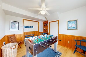 Una habitación con mesa de fútbol, ventilador de techo, paneles de madera, una cesta de mimbre y una alfombra azul.
