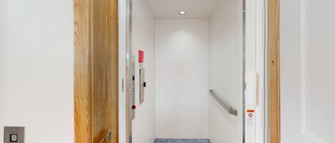 Un pasillo estrecho y bien iluminado, con una puerta de madera a la izquierda, un panel de control y un cartel de prohibido fumar a la derecha.