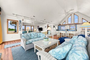 A spacious living area with a high ceiling, a dining table, and a kitchen area with large windows.