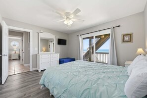 Um quarto com cama, ventilador de teto, televisão, escada para uma varanda e vista para o oceano.