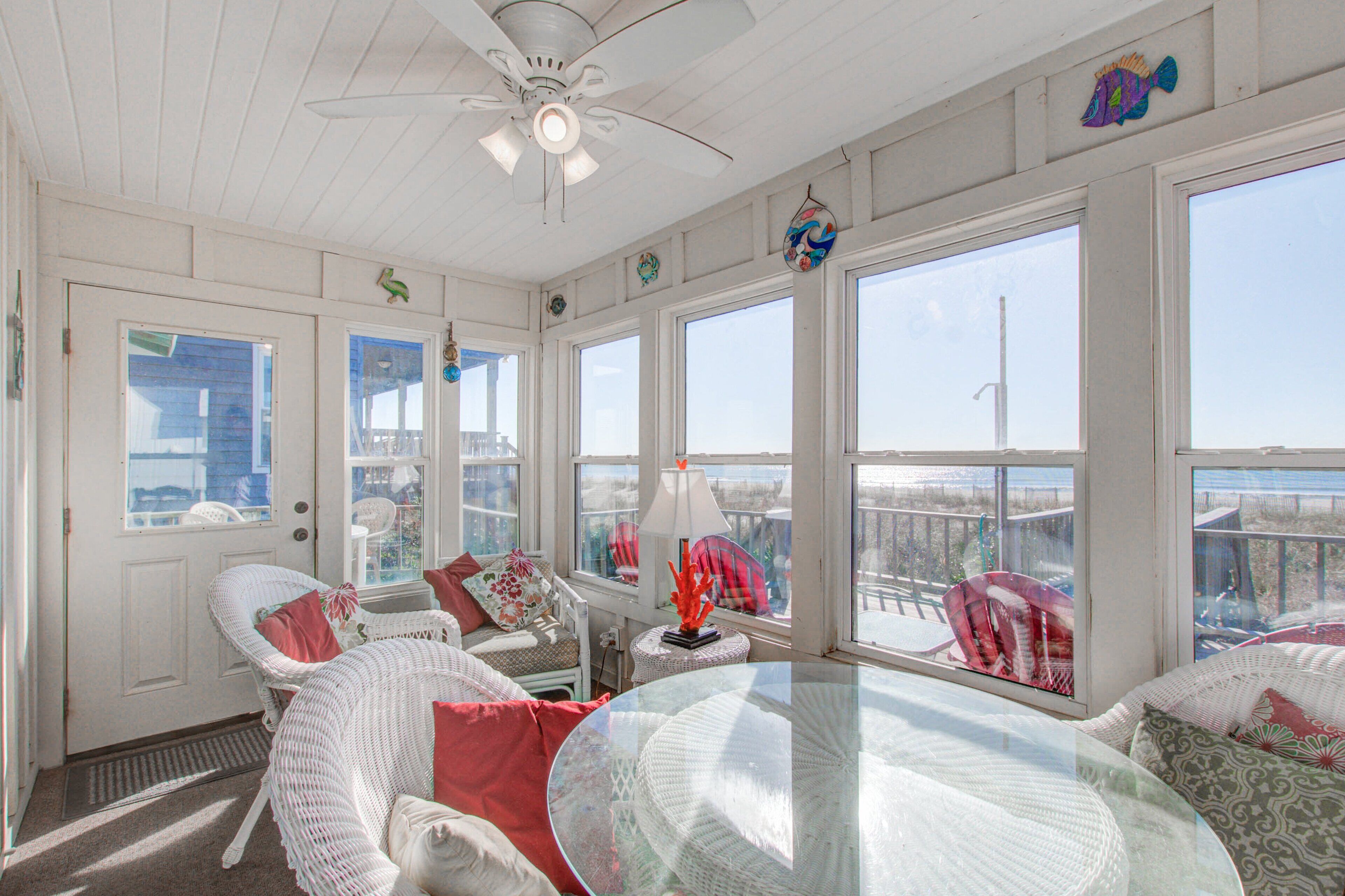 A sunlit room with a glass table, wicker chairs, and large windows offering a view of the beach.