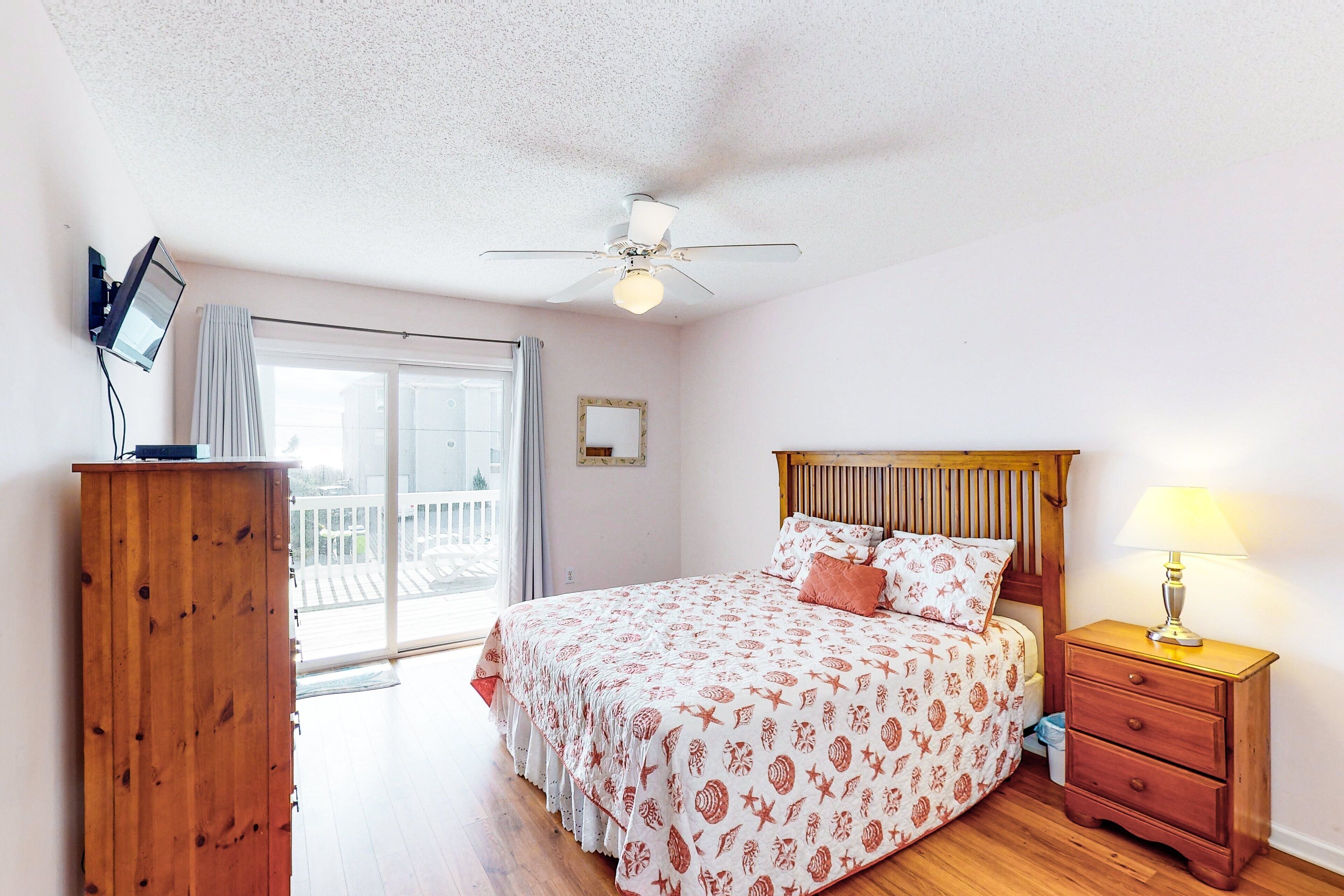A bedroom with a bed, wooden nightstand, ceiling fan, and a view of the outside balcony.