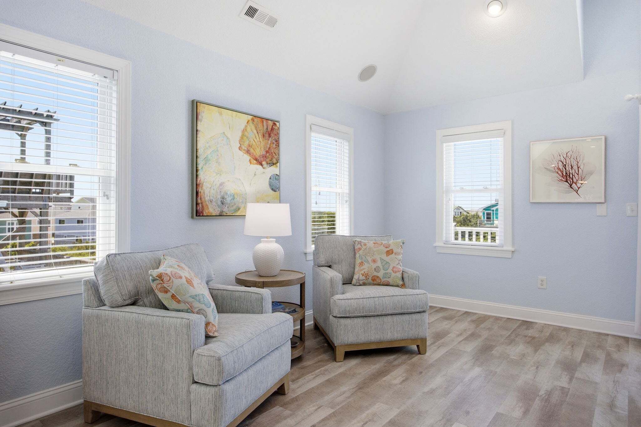 Una habitación con dos sillones grises, una mesita redonda con una lámpara y obras de arte en las paredes.