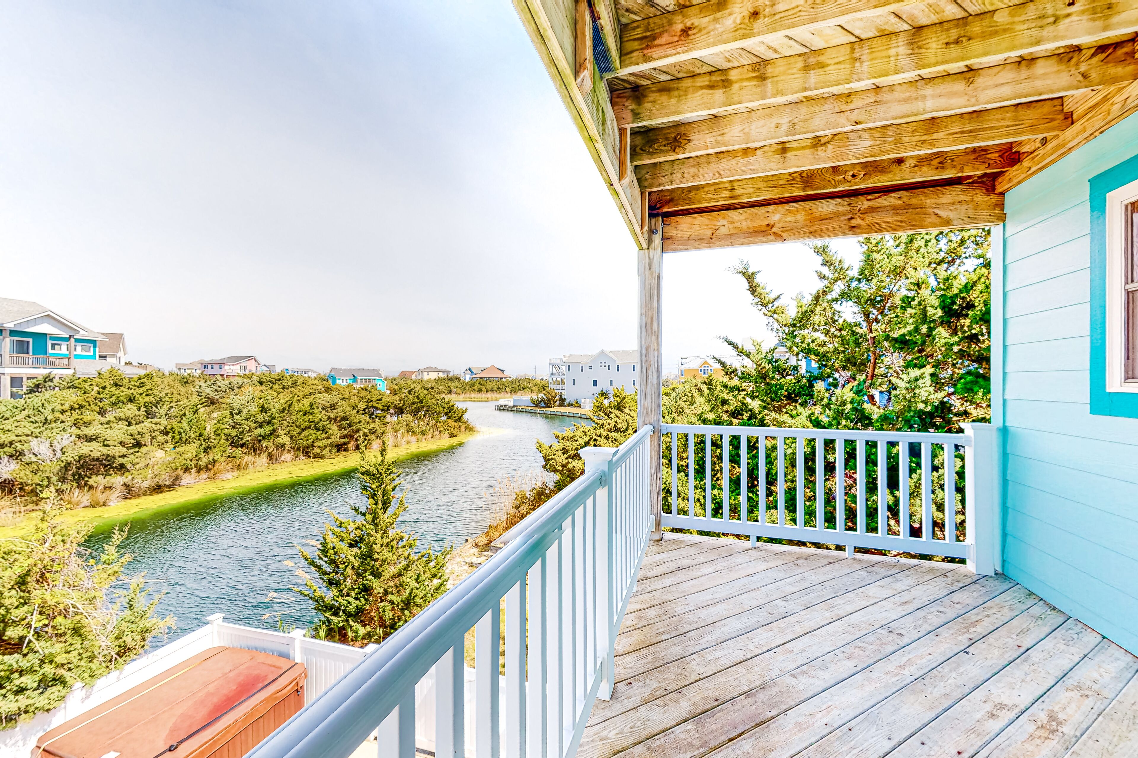 A wooden deck with a railing overlooking a river, surrounded by greenery and residential buildings.