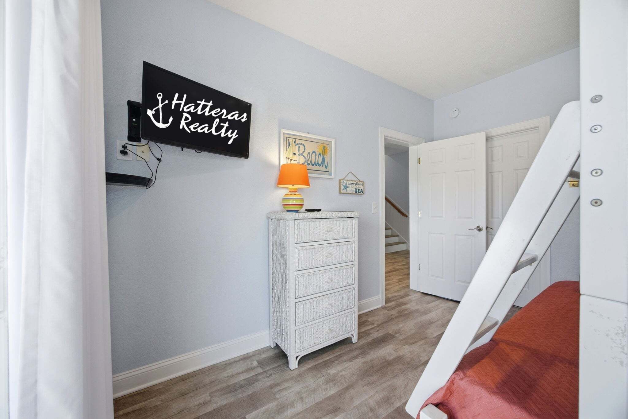 Una stanza con TV a parete che mostra 'Hatteras Realty', un comò bianco in vimini con una lampada e una scala che conduce a un'altra stanza.
