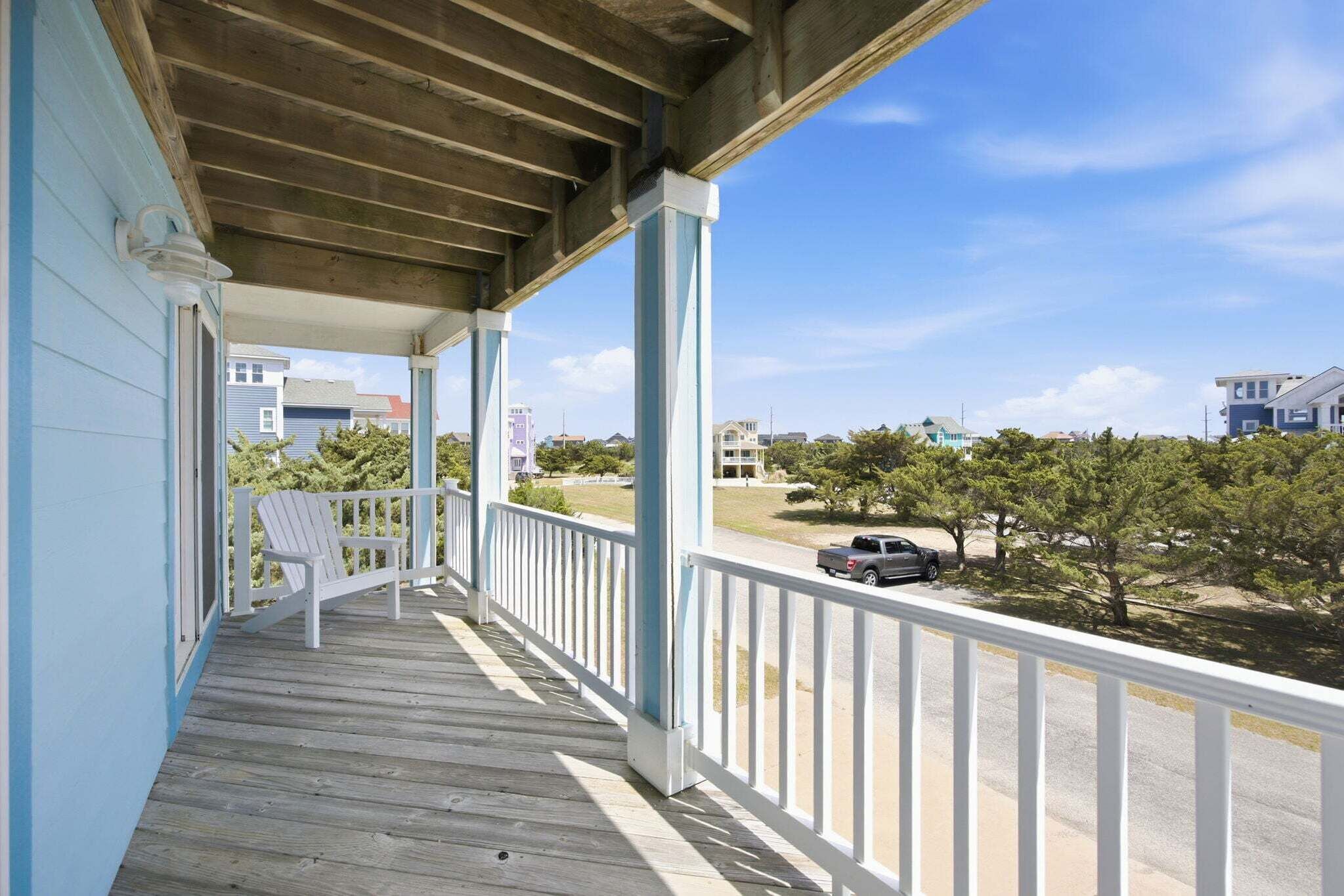 A covered porch with a white railing, a white Adirondack chair, and a view of a street with trees and buildings.