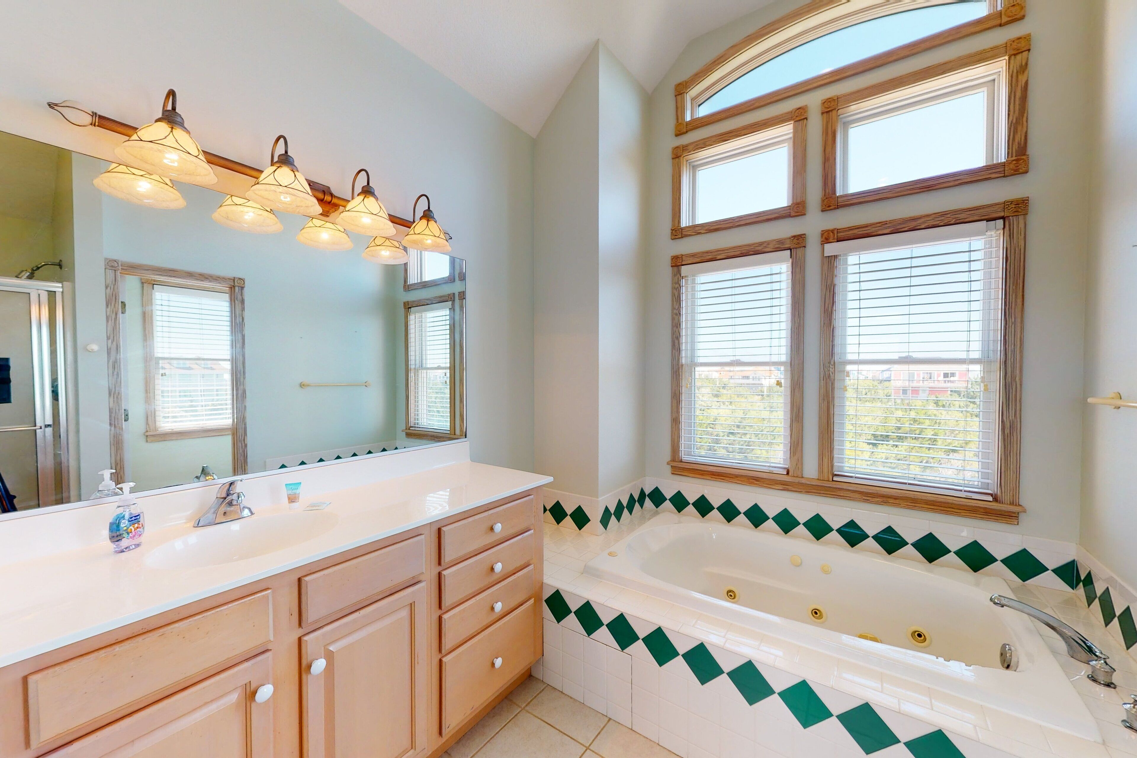 A bathroom with a large mirror, a bathtub with a green and white tile border, and multiple windows with blinds.