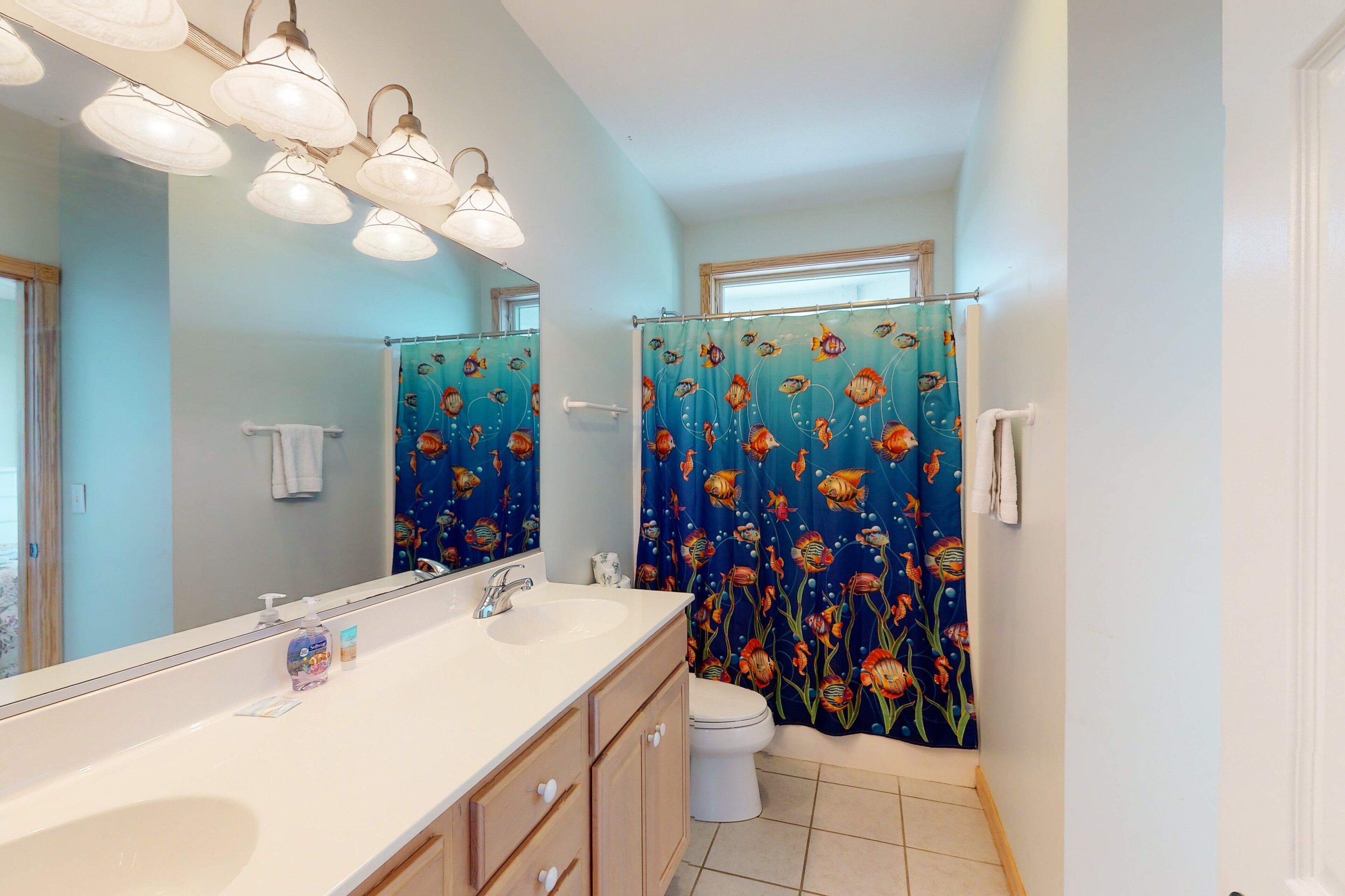 A bathroom with a large mirror, a sink with a soap dispenser, a toilet, and a shower curtain with a fish pattern.