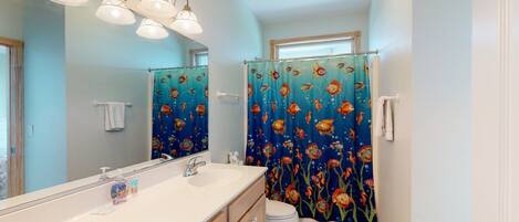 A bathroom with a large mirror, a sink with a soap dispenser, a toilet, and a shower curtain with a fish pattern.