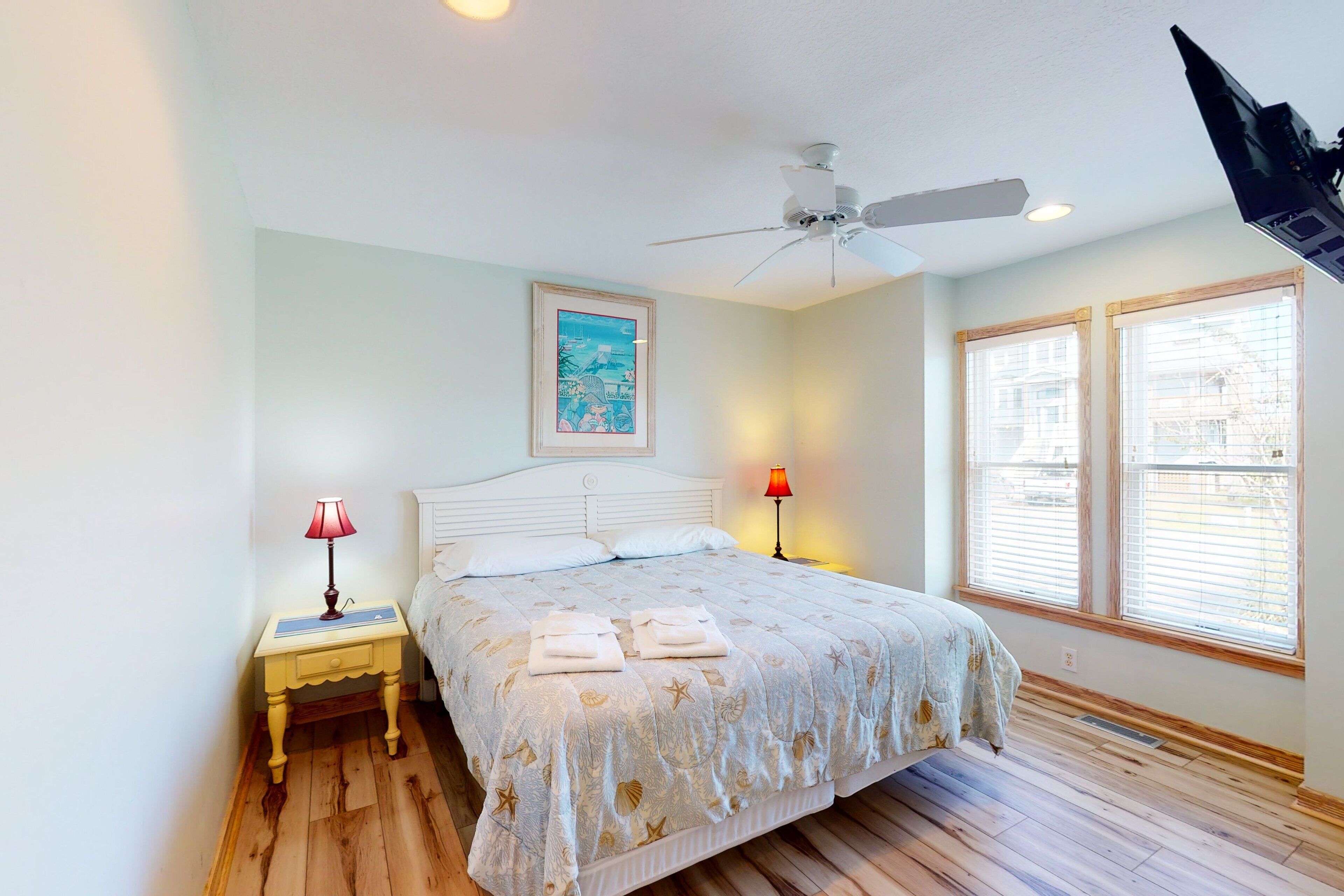 A bedroom with a bed, a ceiling fan, two bedside lamps, and a window with blinds.