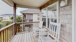 A wooden deck with white chairs and a railing, overlooking a residential area with houses and palm trees. - Dog-friendly duplex in Surf City with beautiful ocean views — just steps from beach access! (Surf City)