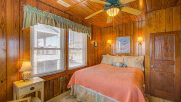 A wooden cabin bedroom with a bed, two bedside lamps, a ceiling fan, and a window with blinds.