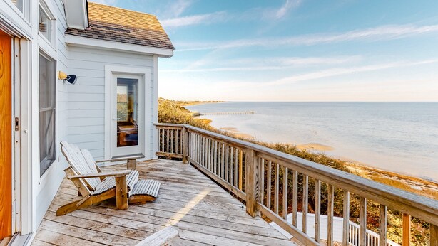 A wooden deck with a white house, a chair, and a view of the ocean. - Waterfront house with private pool, hot tub, W/D, &vocean view (Buxton)
