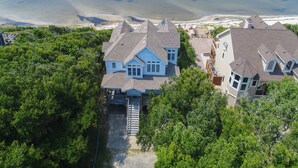 A large, two-story house with a steep roof, surrounded by dense greenery and a beach in the background. - Waterfront house with private pool, hot tub, W/D, &vocean view (Buxton)