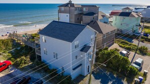 Un quartier résidentiel en bord de mer, avec une grande maison blanche qui se distingue, un garage, une voiture rouge garée devant, et une jetée qui s’avance dans l’océan au loin.