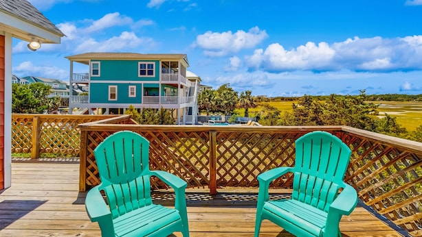 A wooden deck with two green Adirondack chairs, a wooden fence, and a two-story building with a balcony in the background. - HB93 Holden Friends - 2nd row, 3 bedroom, NEW private pool (Holden Beach)