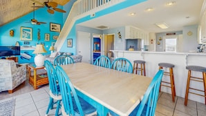 A spacious living room with a wooden ceiling, a dining area with blue chairs, and a kitchen with white cabinets. - HB93 Holden Friends - 2nd row, 3 bedroom, NEW private pool (Holden Beach)