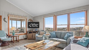 Un vaste salon doté d’une grande fenêtre offrant une vue panoramique, d’un coin salon confortable avec un canapé et des fauteuils, et d’un téléviseur à écran plat fixé au mur.