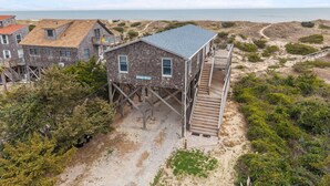 Une maison en bois dotée d’un balcon et d’un escalier menant à une terrasse, entourée de dunes de sable et de végétation.