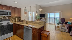 A kitchen with a large island, stainless steel appliances, and wooden cabinets. There is a dining area with a view of the ocean. - Oceanfront condo with beach views, balcony, community pool & W/D, central AC (Surf City)