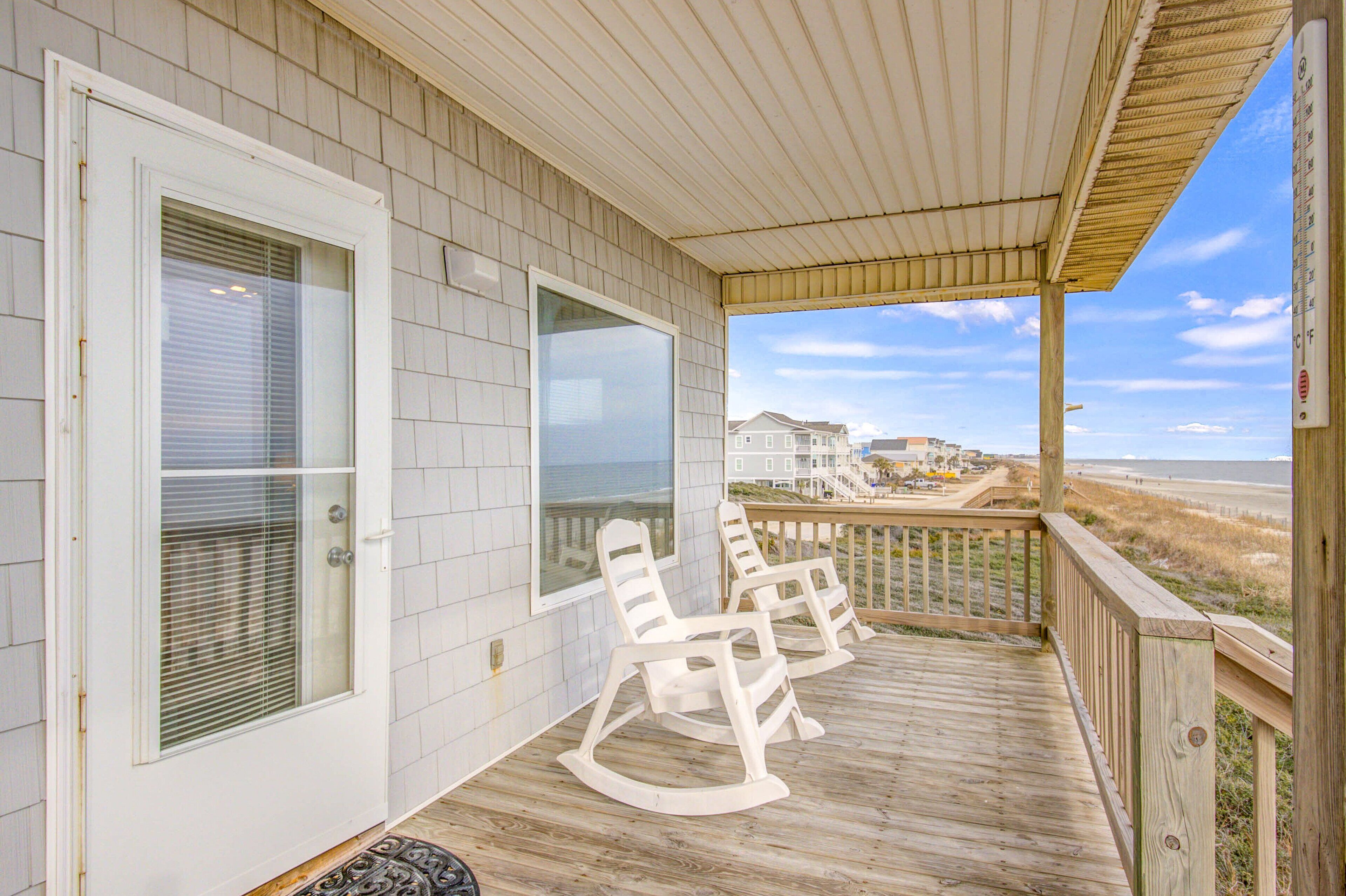 A wooden deck with rocking chairs, a covered porch, and a view of the beach and houses in the background.