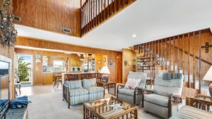 A spacious living area with a high ceiling, wooden walls, and a staircase. There are multiple seating options including a sofa, armchairs, and a coffee table.
