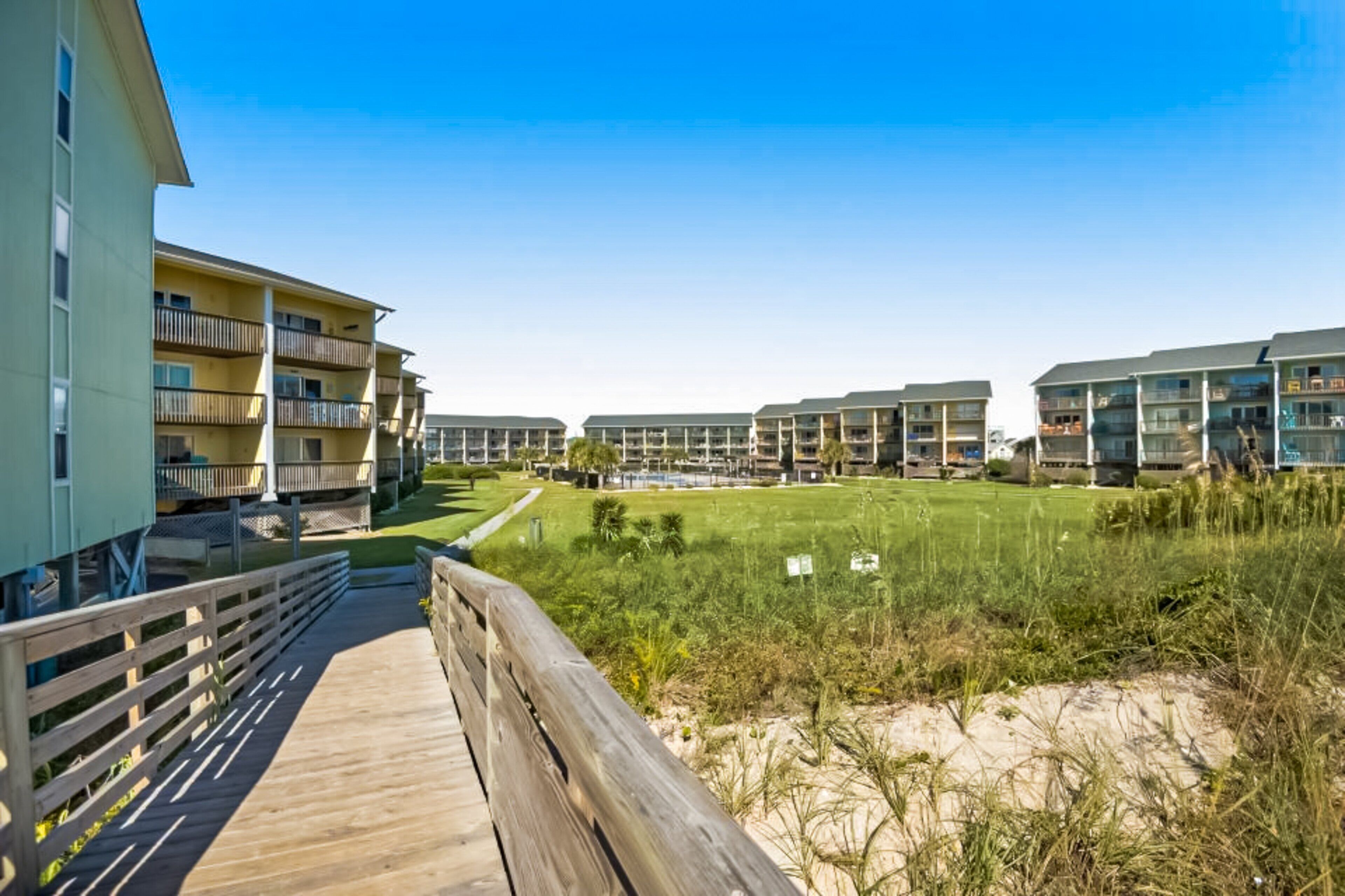 Een strandhotel met meerlaags gebouwen, een houten boardwalk en een grasgebied met duinen.